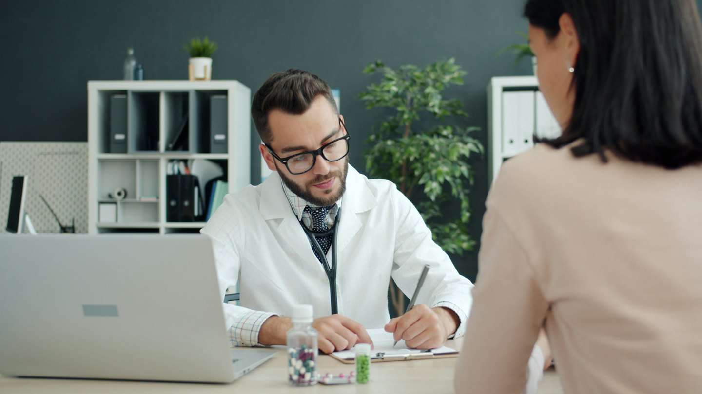 Doctor writes on clipboard during patient consultation at desk.