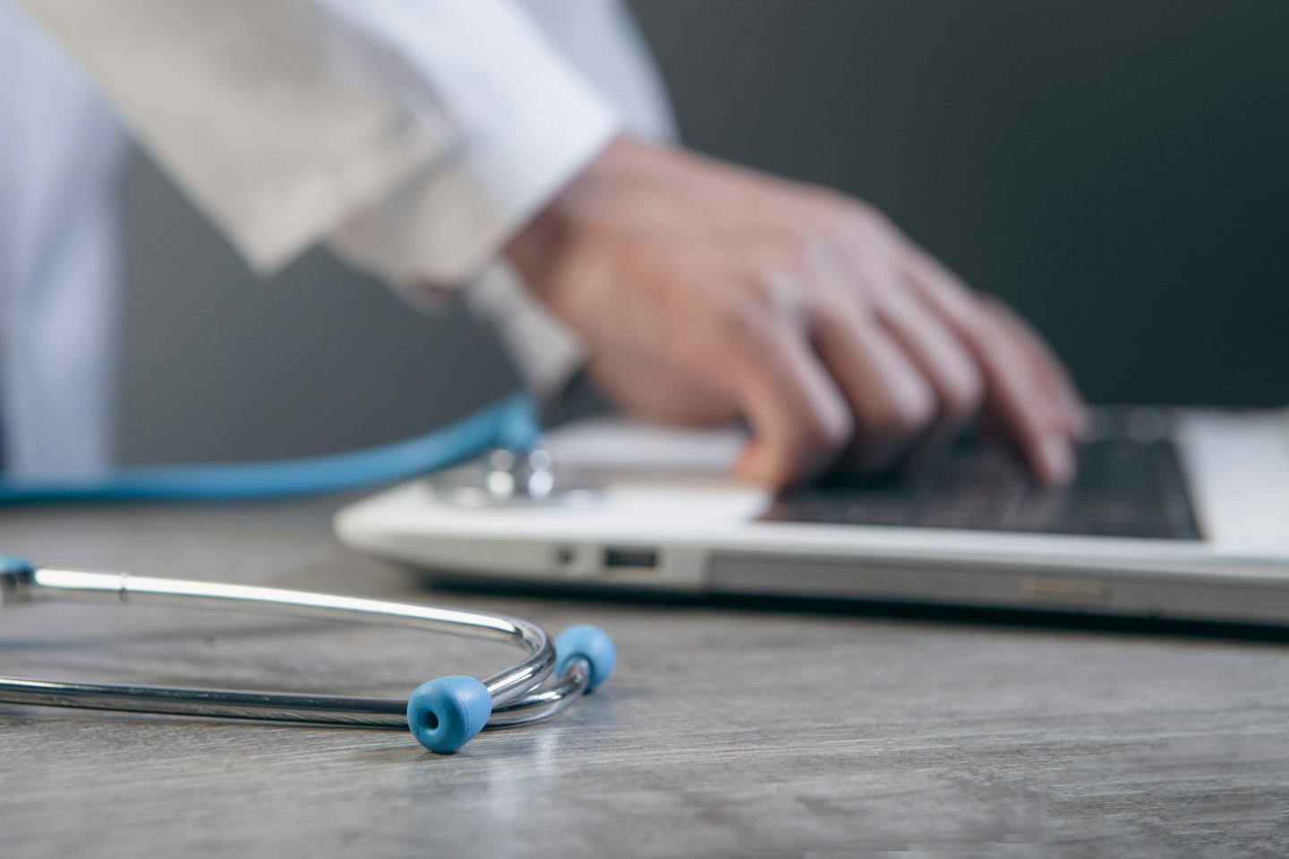 Doctor typing on laptop with stethoscope on desk.